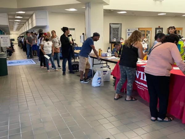 A line of people stand at tables the length of a large room, completing registration for a workshop.